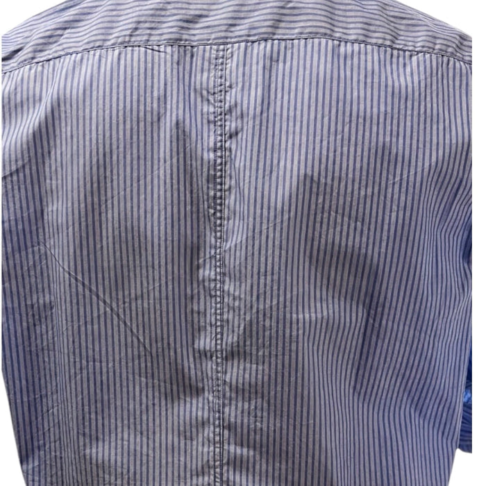 A man in a FRANK & EILEEN Finbar Men's Button Up Shirt in Blue Stripes, blue jeans, and white sneakers walks confidently down a sunny, tree-lined cobblestone street.