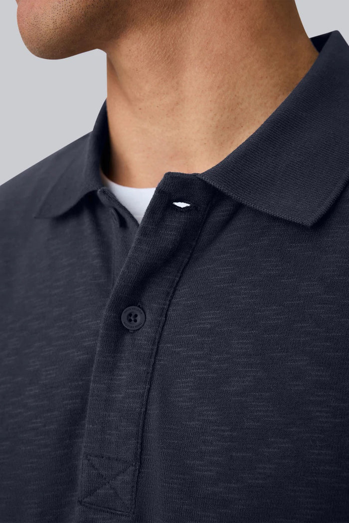 A man with short dark hair is seen from behind, wearing the MACK WELDON - HEAVYWEIGHT SLUB LONG SLEEVE POLO IN FROTH and blue pants, standing against a plain light gray background.