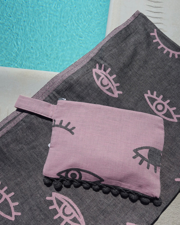 Dark gray towel with pink evil eye print and a pink wet/dry zippered pouch clytch bag on top, next to a pool