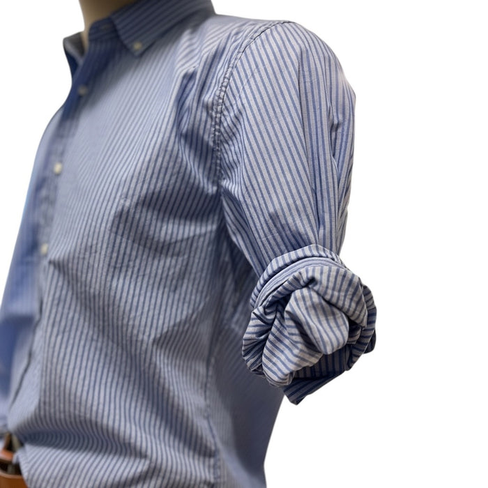 A man in a FRANK & EILEEN Finbar Men's Button Up Shirt in Blue Stripes, blue jeans, and white sneakers walks confidently down a sunny, tree-lined cobblestone street.