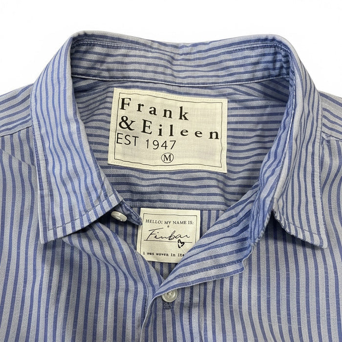 A man in a FRANK & EILEEN Finbar Men's Button Up Shirt in Blue Stripes, blue jeans, and white sneakers walks confidently down a sunny, tree-lined cobblestone street.