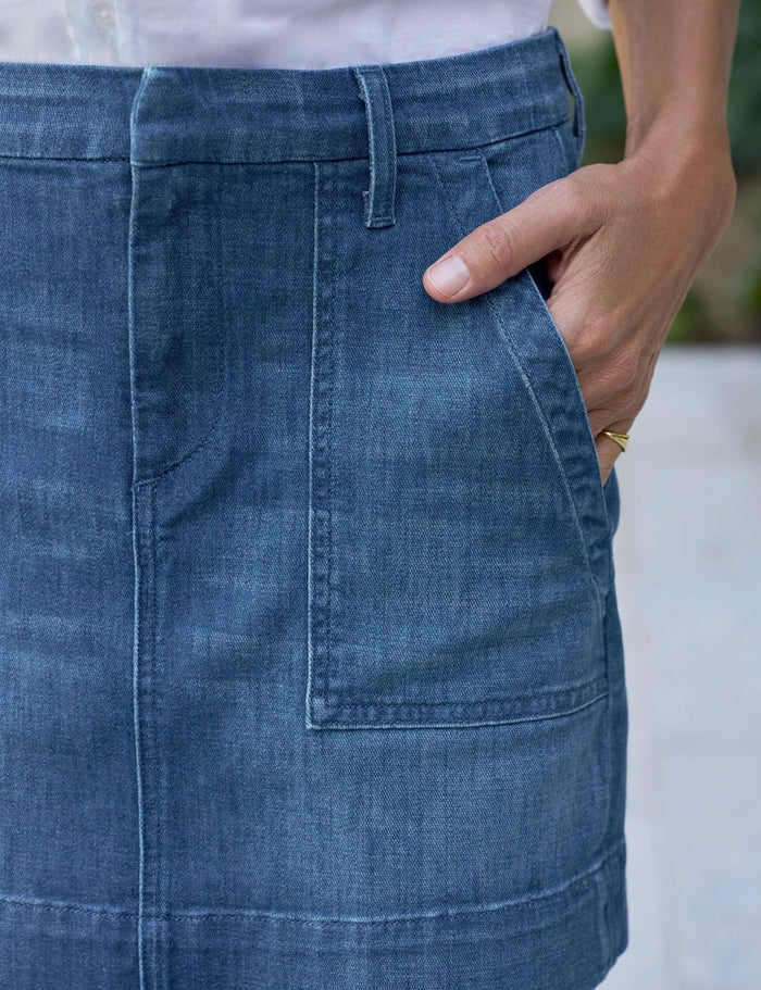 Close-up of a person wearing the FRANK & EILEEN Jameson Utility Skirt in Italian Dream Light Denim 1984 Wash, with utility pockets, a white shirt, and a gold ring. Their right hand rests in the front pocket; background is blurred.