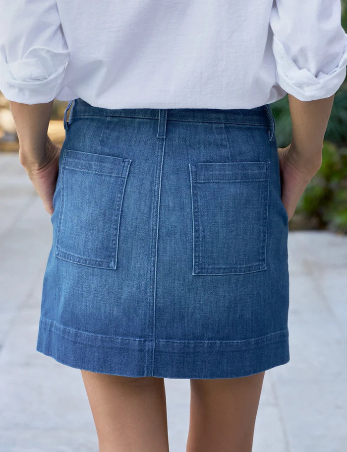 Close-up of a person wearing the FRANK & EILEEN Jameson Utility Skirt in Italian Dream Light Denim 1984 Wash, with utility pockets, a white shirt, and a gold ring. Their right hand rests in the front pocket; background is blurred.