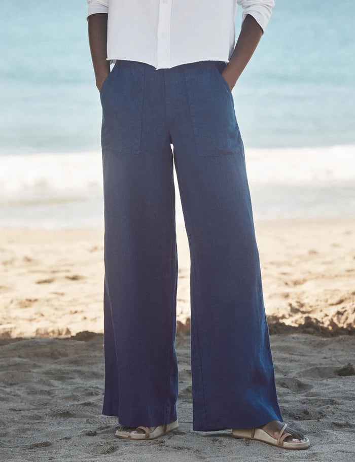 A person stands on a sandy beach wearing a white long-sleeve shirt, FRANK & EILEEN MADELINE Patch Pocket Pull On Wide Leg Linen Pant in Navy, and light-colored sandals. The ocean is visible in the background.