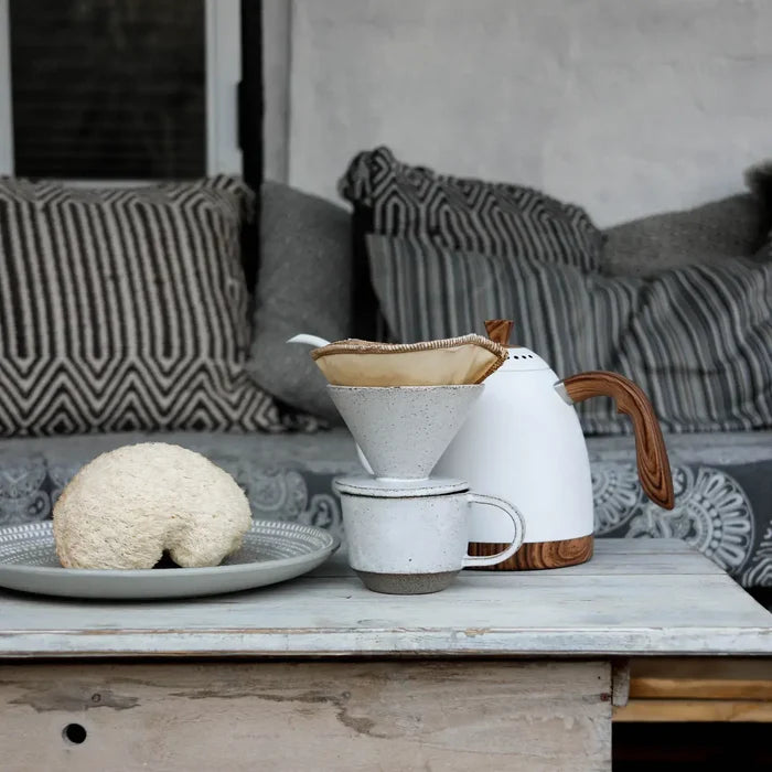 White ceramic coffee maker with wooden handle on a wooden table, with a sofa and cushions in the background.