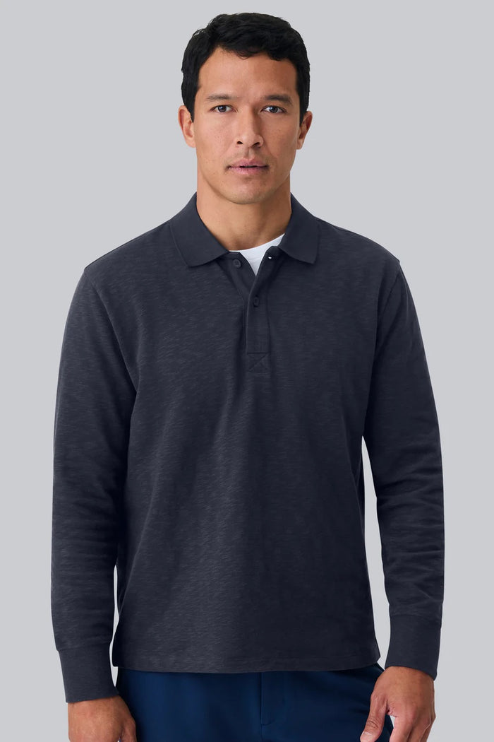A man with short dark hair is seen from behind, wearing the MACK WELDON - HEAVYWEIGHT SLUB LONG SLEEVE POLO IN FROTH and blue pants, standing against a plain light gray background.