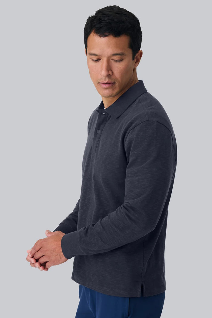 A man with short dark hair is seen from behind, wearing the MACK WELDON - HEAVYWEIGHT SLUB LONG SLEEVE POLO IN FROTH and blue pants, standing against a plain light gray background.