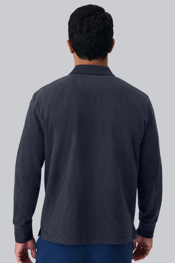 A man with short dark hair is seen from behind, wearing the MACK WELDON - HEAVYWEIGHT SLUB LONG SLEEVE POLO IN FROTH and blue pants, standing against a plain light gray background.