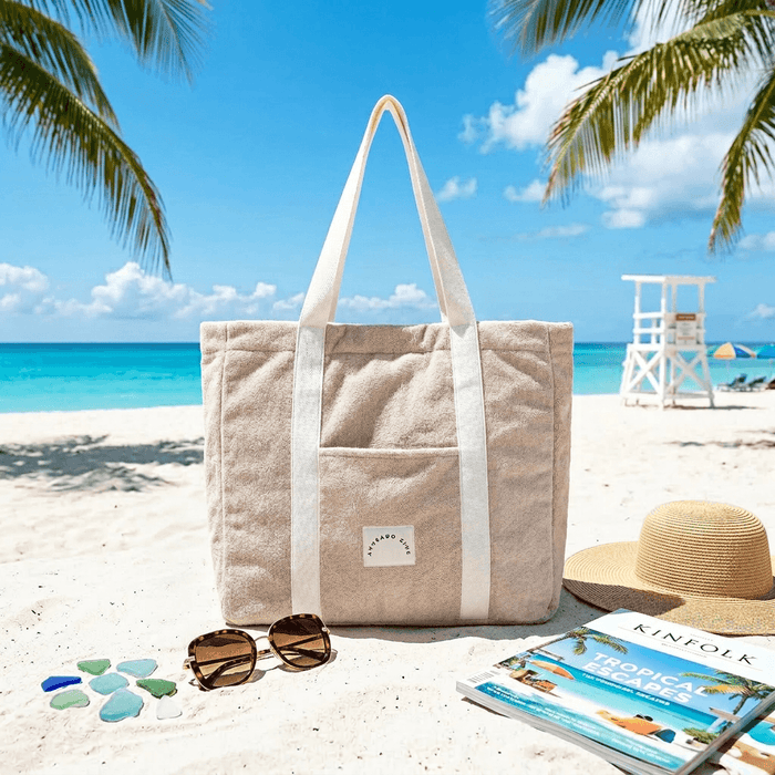 Beige tote bag on a sandy beach with sunglasses, a hat, and a book.