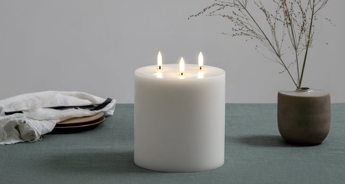 The PIFFANY UYUNI - IVORY TRIPLE FLAME PILLAR CANDLE 6X5.9 features three lit wicks that burn evenly at the top center of the large, ivory-colored candle, set against a plain white background.